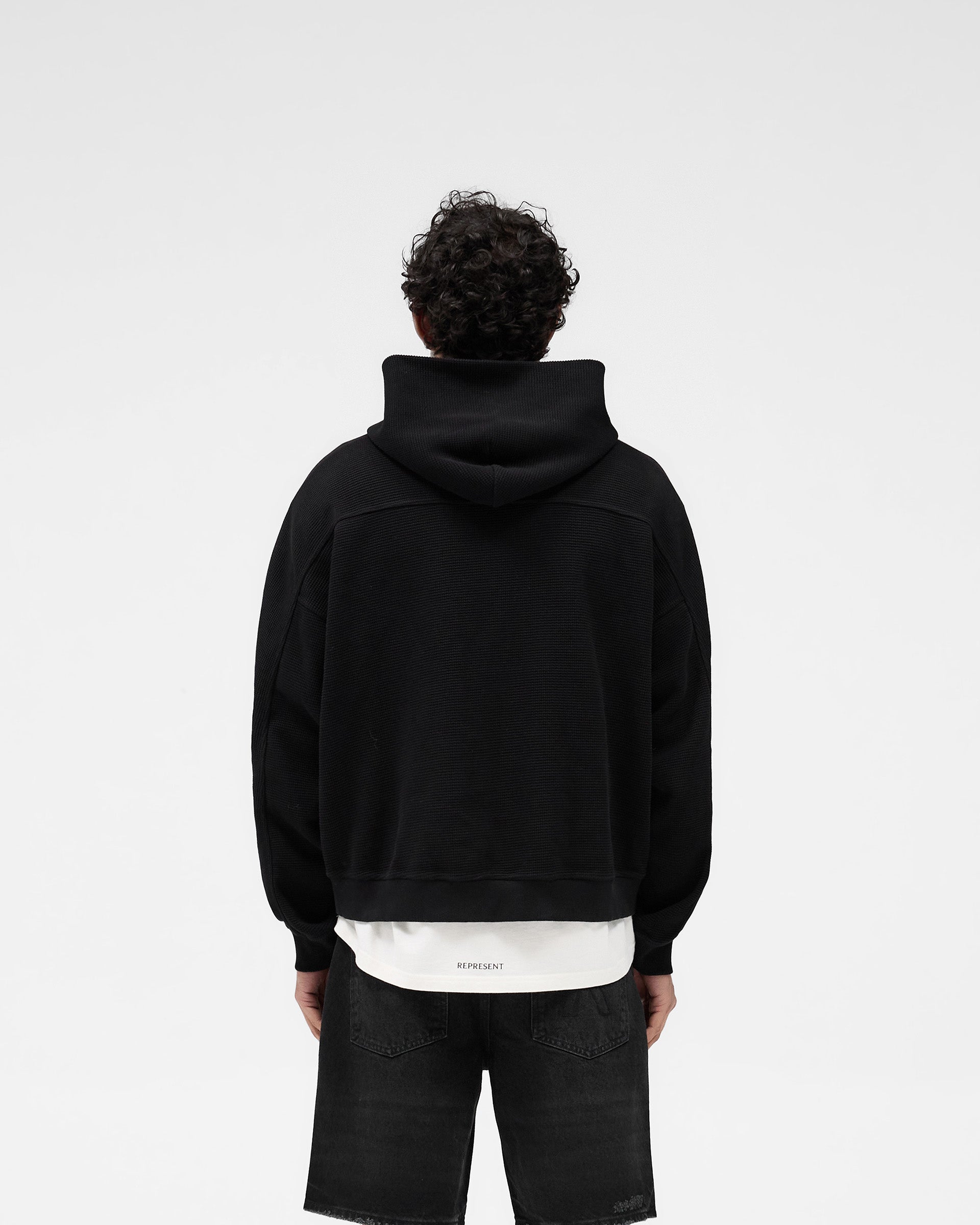 Man wearing an oversized black hoodie on a plain white background | Waffle Zip Through Hoodie in Jet Black | Boxy fit hoodie with embroidered logo and RiRi zipper | SS25 Hoodies | Represent Clo