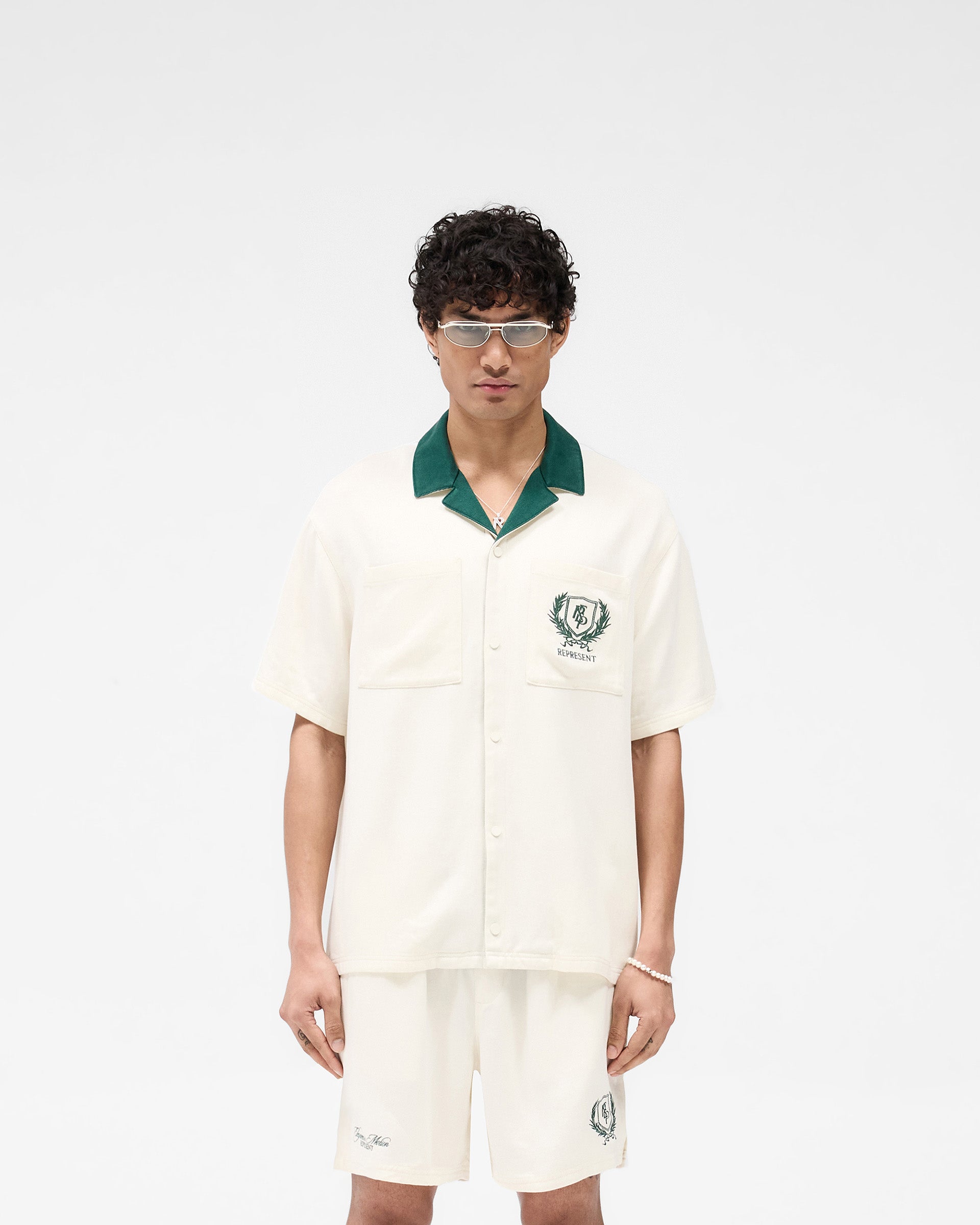 Man wearing an antique white short sleeve shirt with a forest green collar on a plain white background | Represent Crest Shirt in Antique White | Short sleeve shirt with dual chest pockets and embroidered back graphic | SS25 Shirts | Represent Clo